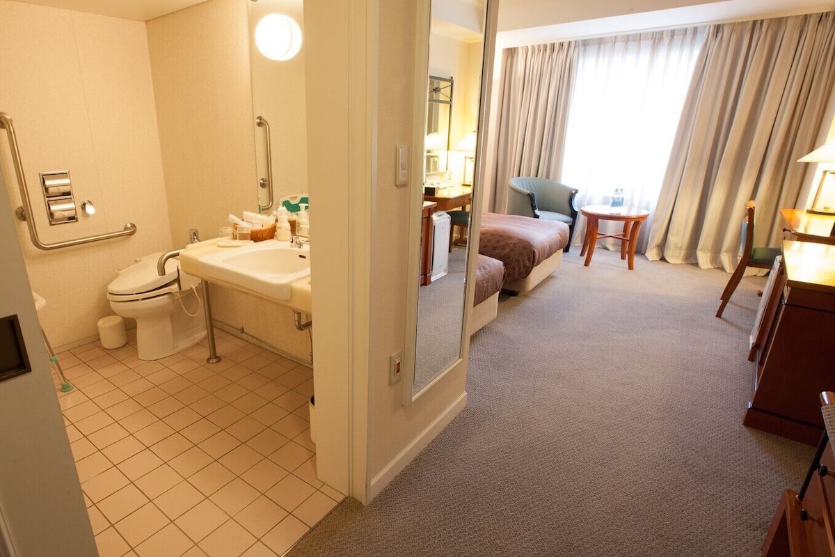 Ein Hotelbad mit Toilette, Waschbecken und Spiegel. Im angrenzenden Zimmer befindet sich ein Bett und ein kleiner Tisch mit einer Lampe.