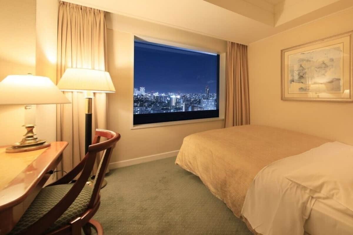 Ein Hotelzimmer mit Bett, Schreibtisch, Stuhl, Fenster mit Blick auf die Stadt und einem gerahmten Bild an der Wand.
