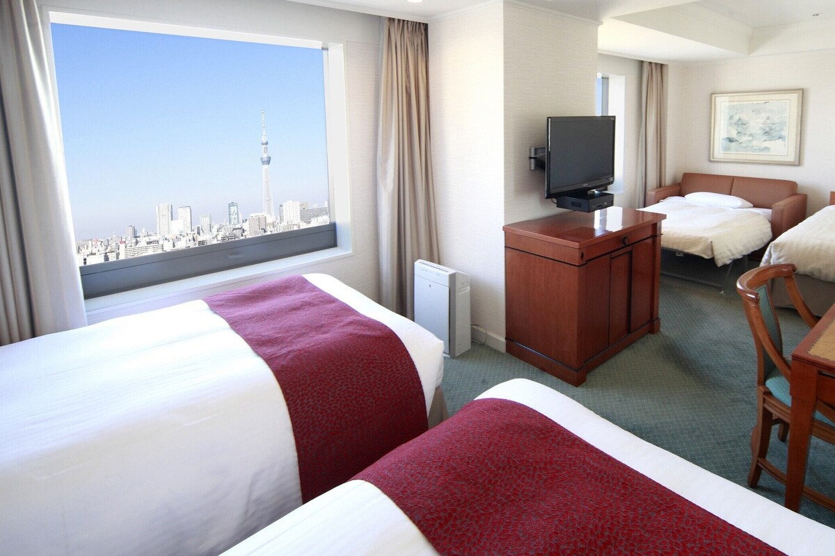 Una habitación de hotel con dos camas, un escritorio, un televisor y una ventana con vista a la ciudad.