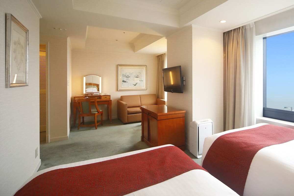Ein Hotelzimmer mit zwei Betten, einem Schreibtisch mit Stuhl, einem Fernseher und einem großen Fenster mit Blick auf die Stadt.