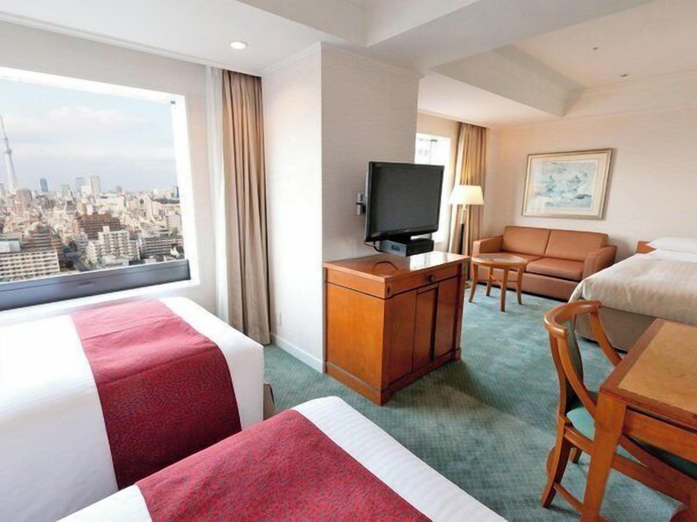 A hotel room with two beds, a TV, a sofa, a desk, and a view of a cityscape.