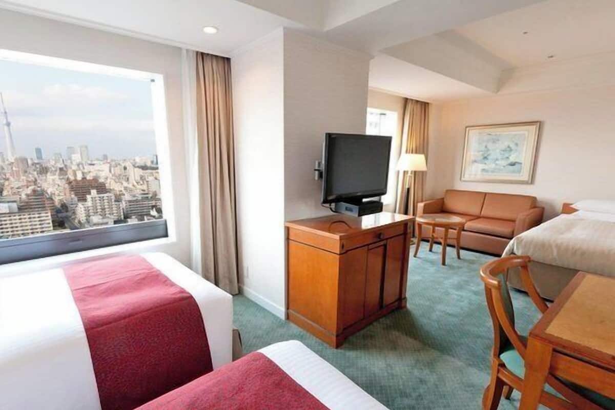 Ein Hotelzimmer mit zwei Betten, einem Fernseher, einem Sofa, einem Schreibtisch und Blick auf eine Stadtlandschaft.