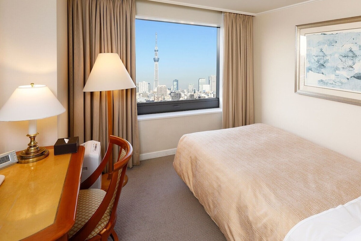 Ein Hotelzimmer mit Bett, Schreibtisch mit Lampe, Stuhl, Fenster mit Blick auf die Stadt und einem gerahmten Bild an der Wand.