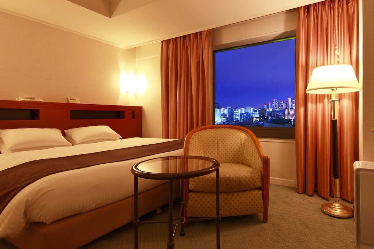 Ein Hotelzimmer mit Bett, Sessel, kleinem Tisch, Lampe und Blick auf eine nächtliche Skyline.