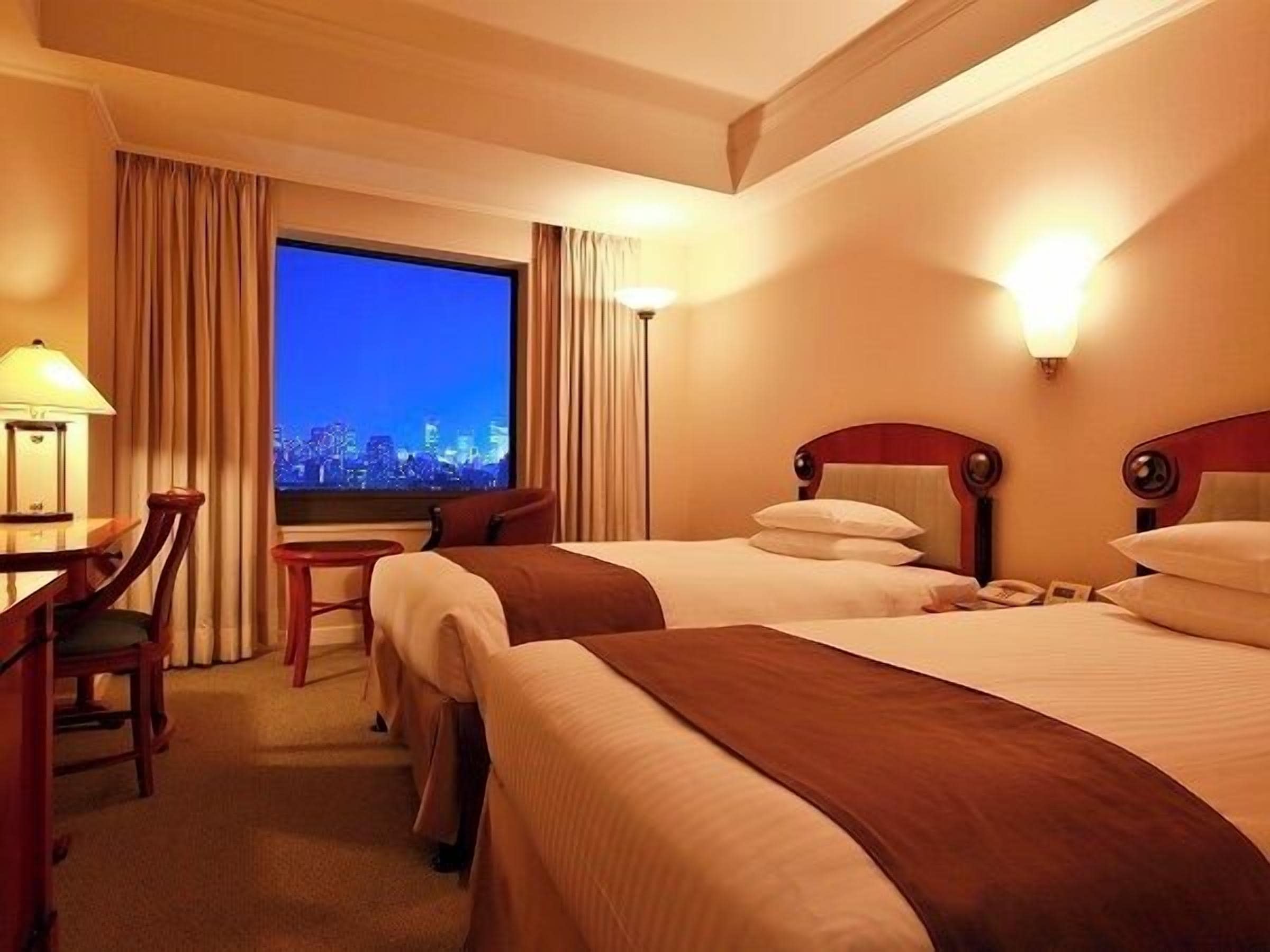 A hotel room with two beds, a desk, a chair, a lamp, and a window with a city view.