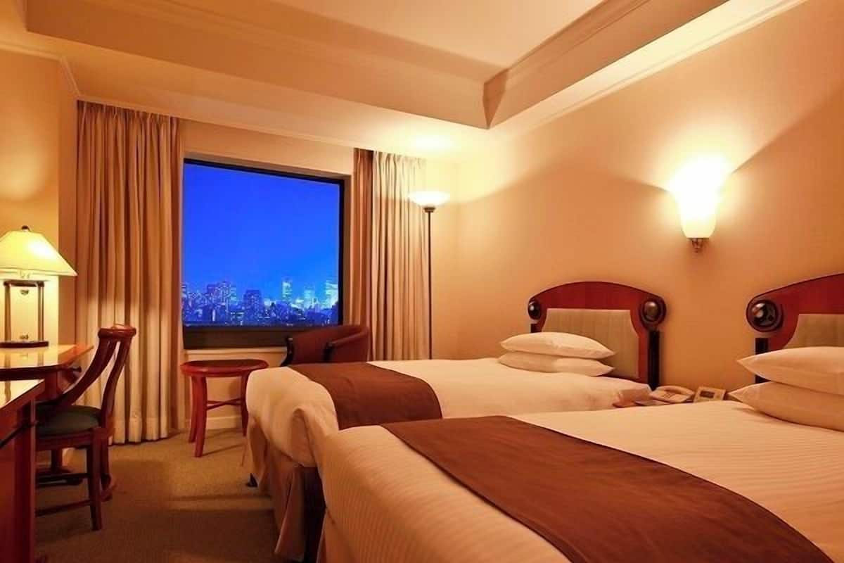 Ein Hotelzimmer mit zwei Betten, einem Schreibtisch, einem Stuhl, einer Lampe und einem Fenster mit Blick auf die Stadt.