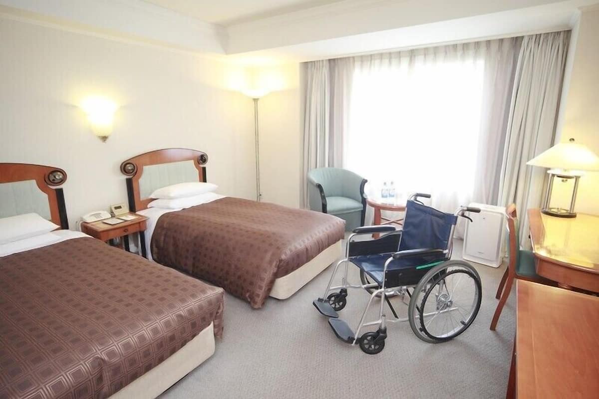 Ein Hotelzimmer mit zwei Betten, einem Rollstuhl, einem Schreibtisch und einem Stuhl.