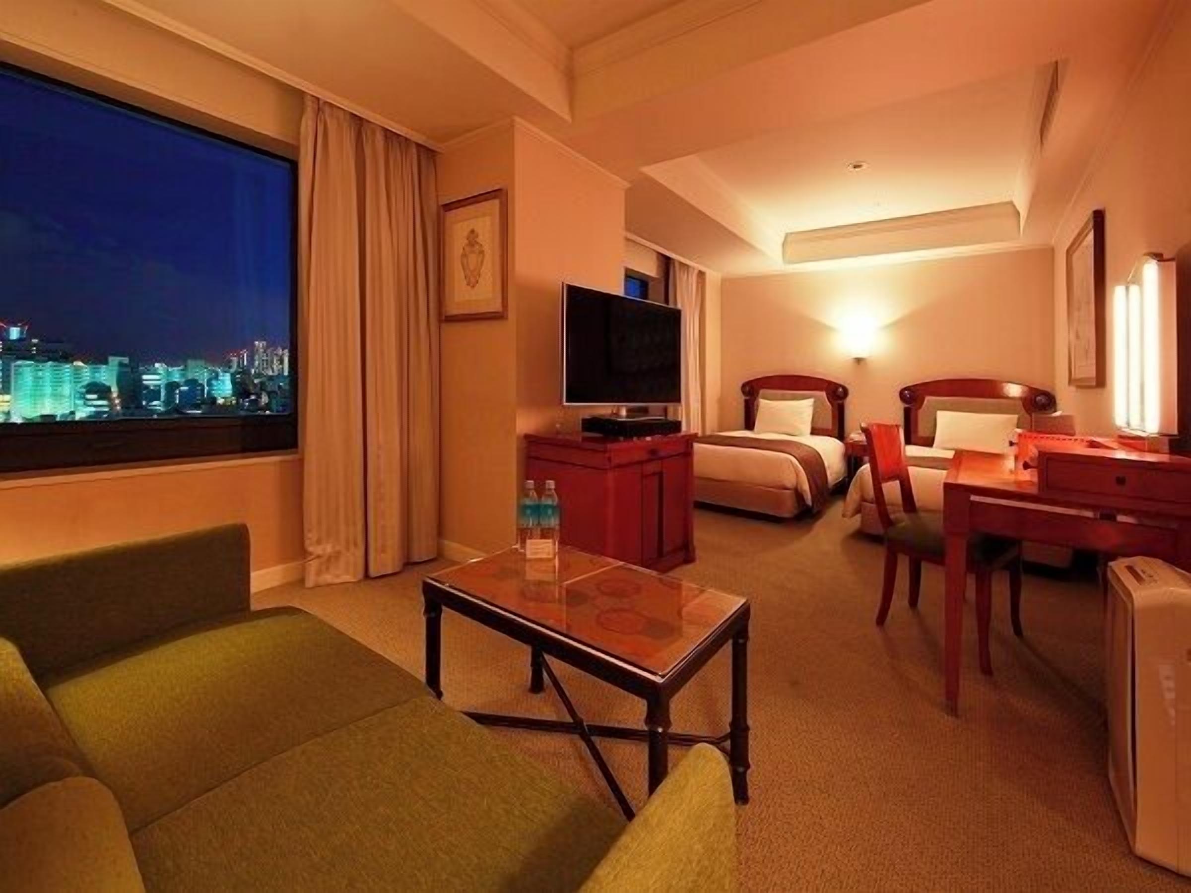 A hotel room with a bed, a desk, a television, a window with a city view, and a chair.