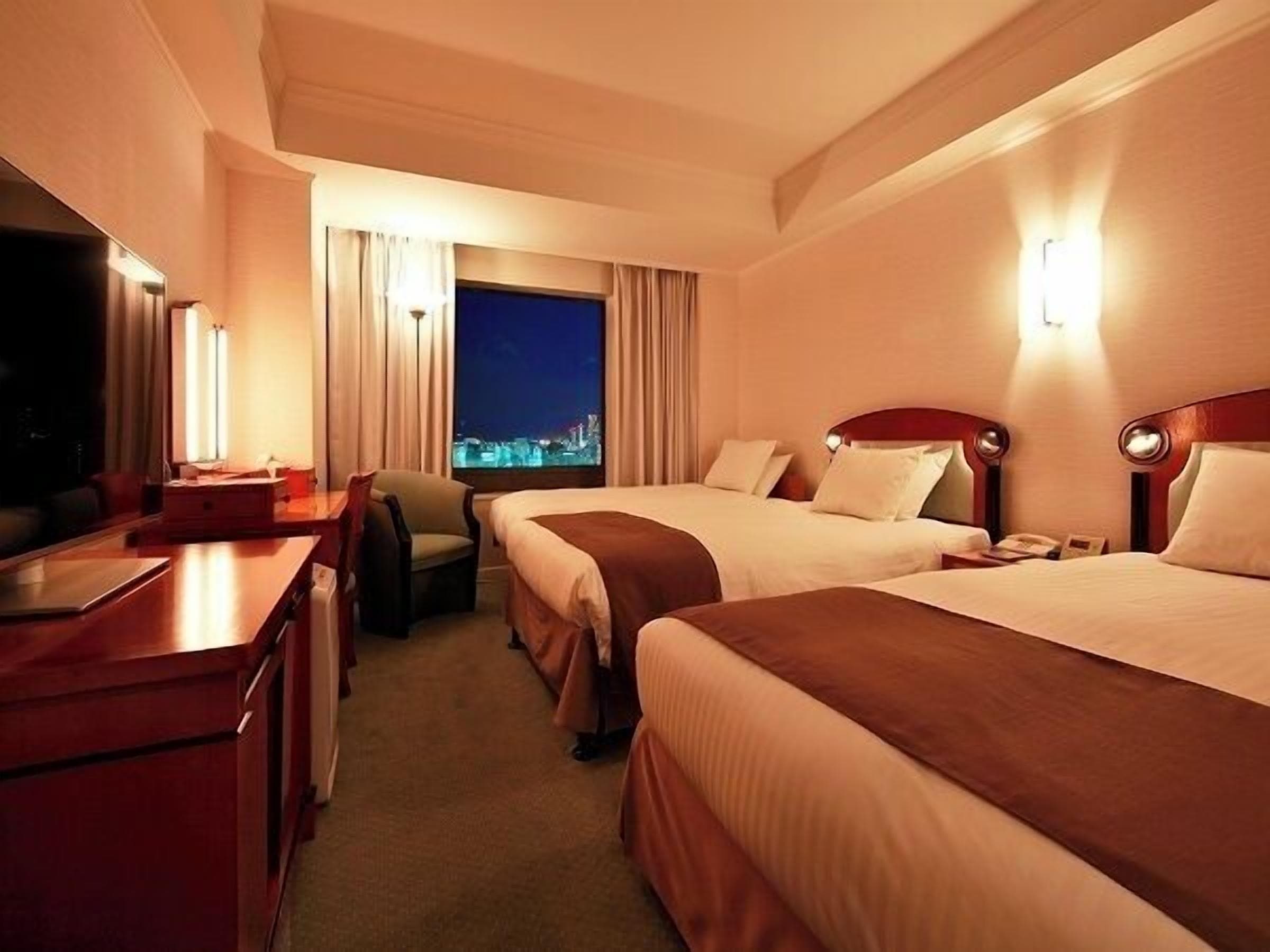 A hotel room with two beds, a desk, a chair, and a large window with a city view.