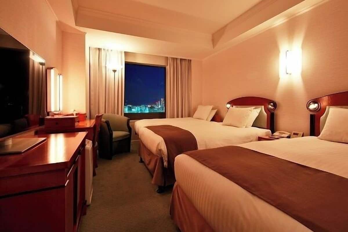 Ein Hotelzimmer mit zwei Betten, einem Schreibtisch, einem Stuhl und einem großen Fenster mit Blick auf die Stadt.