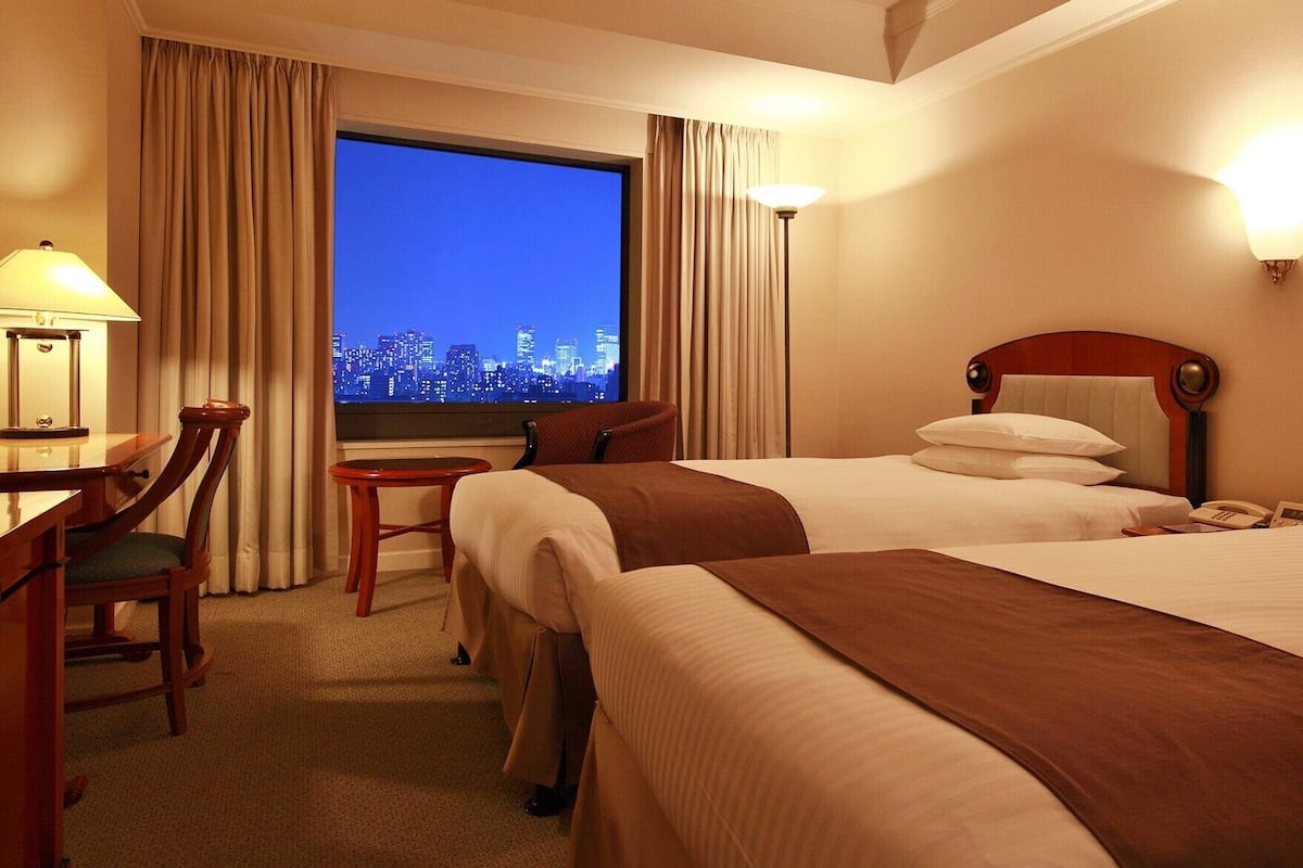 Ein Hotelzimmer mit zwei Betten, einem Schreibtisch, einem Stuhl, einer Lampe und Blick auf eine nächtliche Stadtansicht.