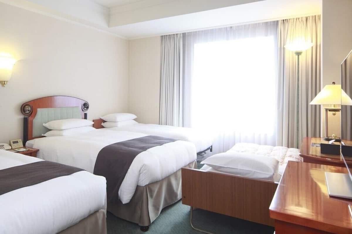 Ein Hotelzimmer mit zwei Betten, einem Schreibtisch, einem Stuhl, einer Lampe und einem großen Fenster mit Vorhängen.