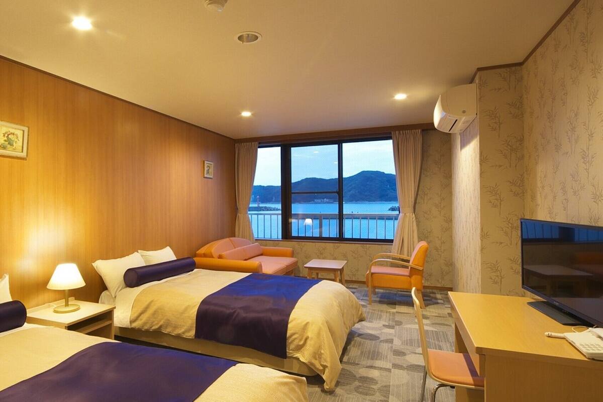 A hotel room with two beds, a desk, a chair, a TV, and a large window with a view of the sea.