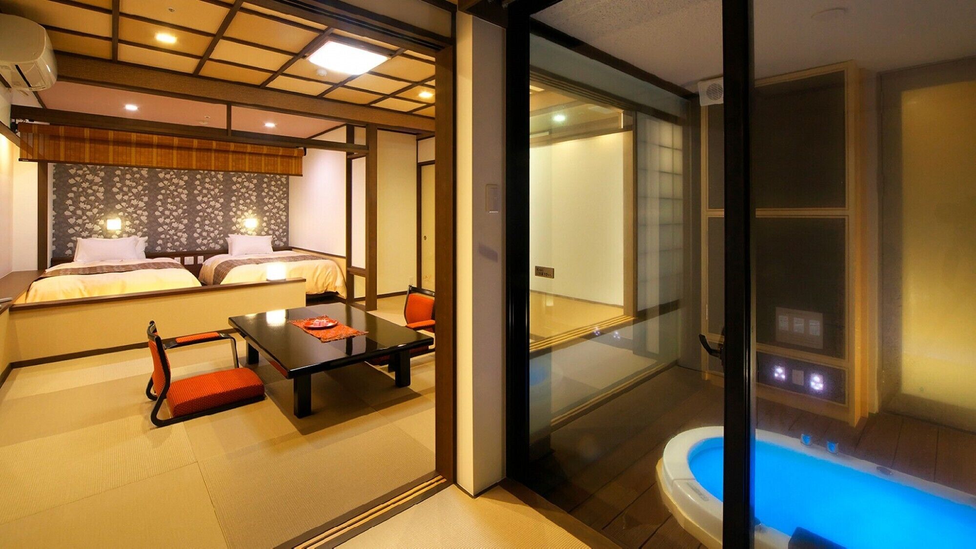 A hotel room with a bed, a small table, a chair, a glass-enclosed bathroom with a hot tub, and tatami flooring.