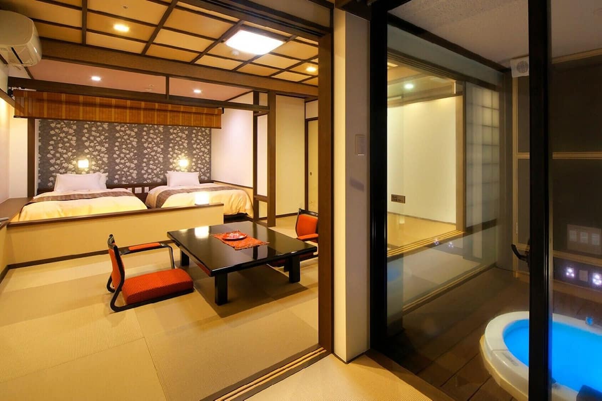 A hotel room with a bed, a small table, a chair, a glass-enclosed bathroom with a hot tub, and tatami flooring.