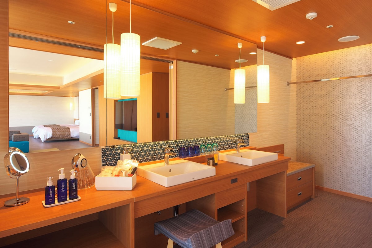 A hotel bathroom with two sinks, a large mirror, and a bench.