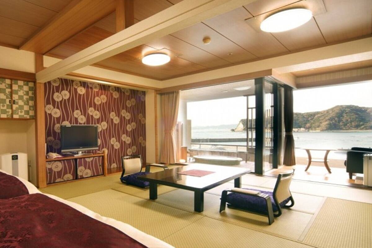 A room with a bed, a TV, a dining table, and a view of the sea.