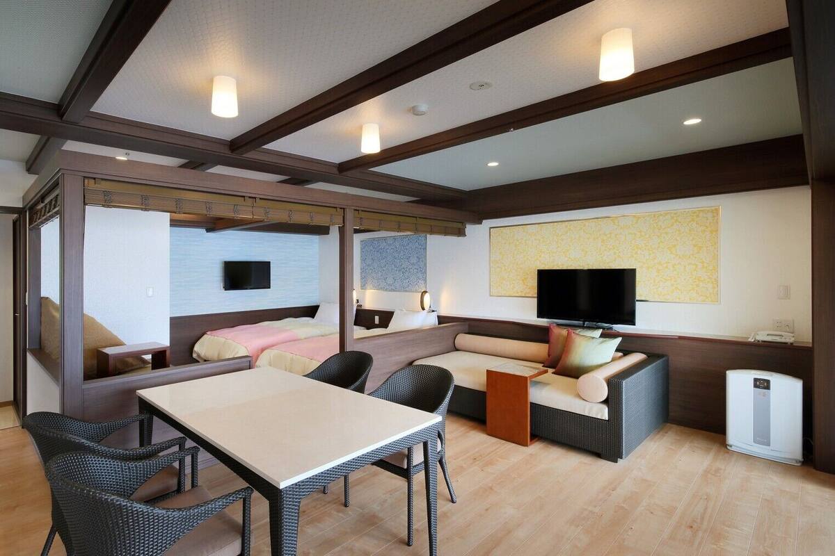 A hotel room with a bed, sofa, dining table, chairs, TV, and a heater.