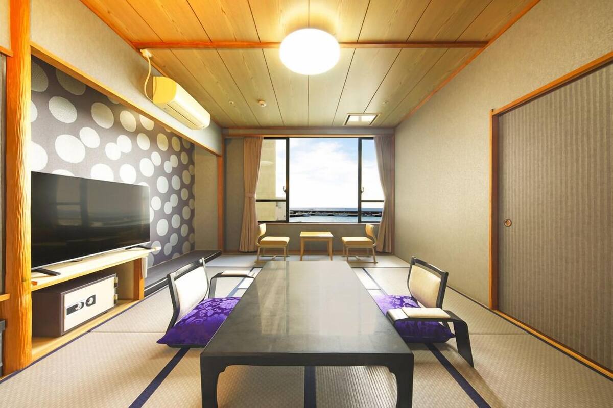 A room with a large flat-screen TV, a long table with chairs, a view through large windows, and a traditional Japanese-style wall with circular patterns.