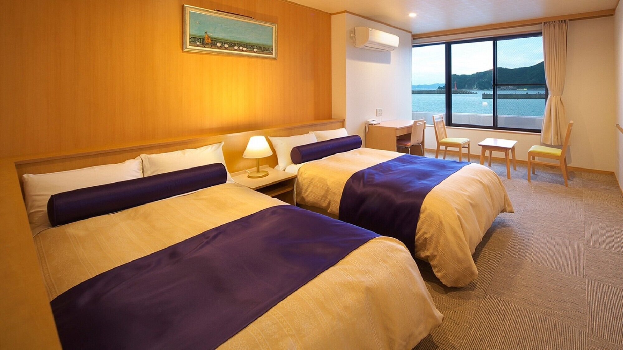 A hotel room with two beds, a desk, a chair, and a large window with a view of the water.
