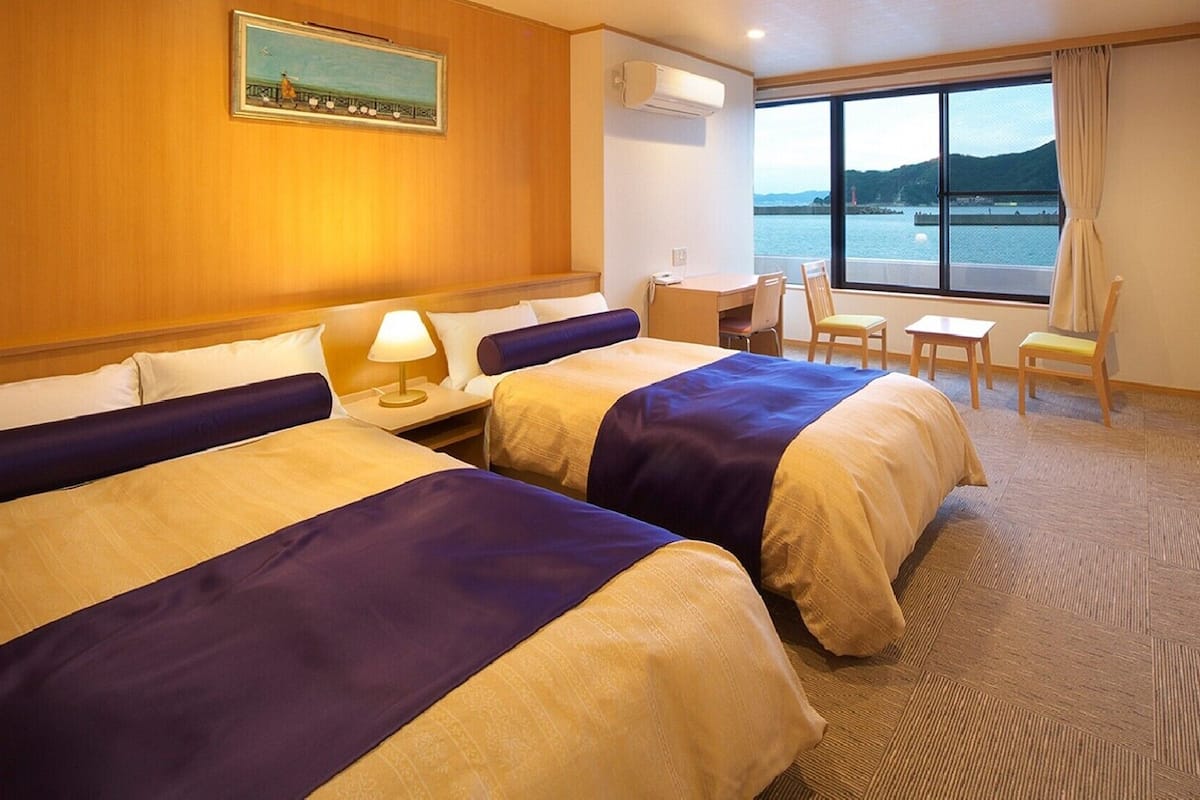 Ein Hotelzimmer mit zwei Betten, einem Schreibtisch, einem Sessel und einem großen Fenster mit Blick auf das Wasser.
