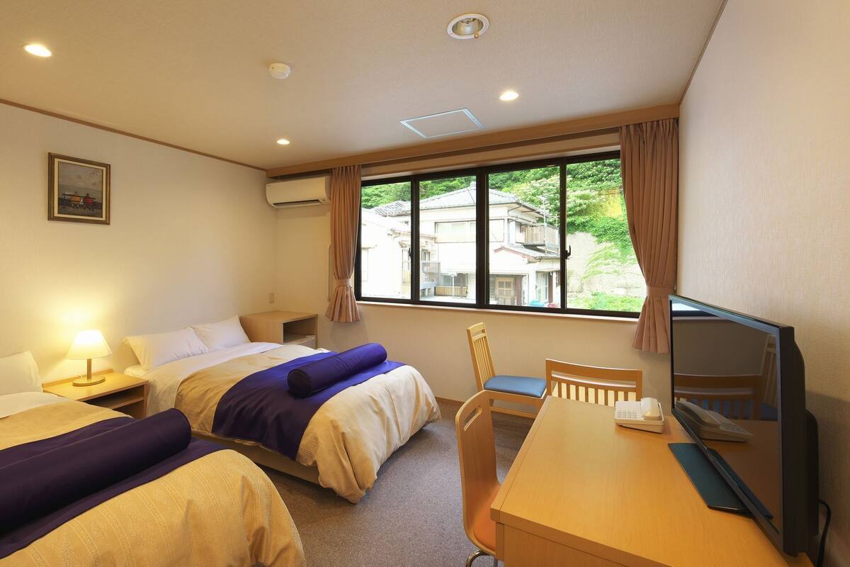 A hotel room with two beds, a desk with a computer, a chair, and a window with a view of buildings.