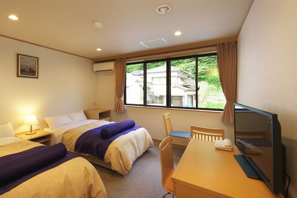 Ein Hotelzimmer mit zwei Betten, einem Schreibtisch mit Computer, einem Stuhl und einem Fenster mit Blick auf Gebäude.