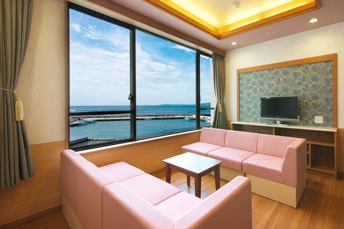 A room with a large window, a pink sofa, a small table, and a TV on a stand.