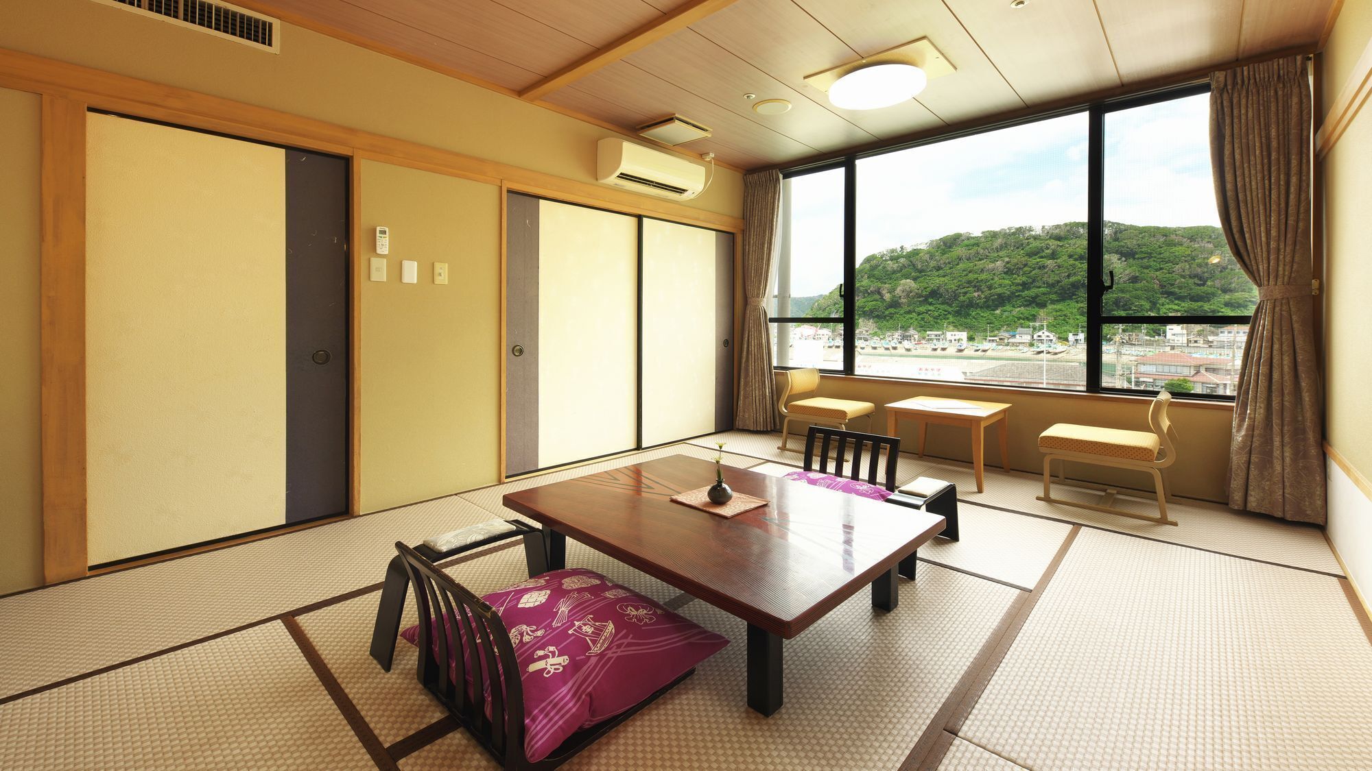 A traditional Japanese room with tatami flooring, a wooden table, chairs, and a view of a green landscape through large windows.