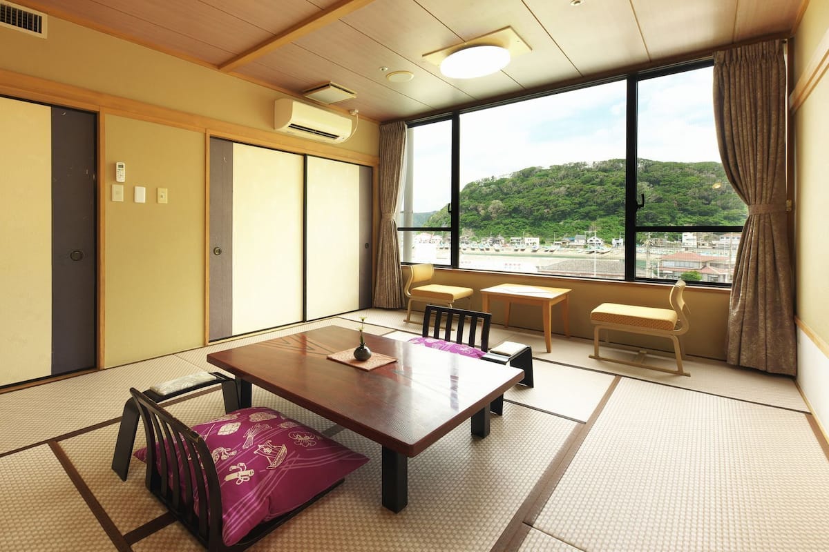 A traditional Japanese room with tatami flooring, a wooden table, chairs, and a view of a green landscape through large windows.