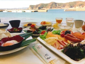 Une table dressée avec divers plats, notamment des sushis, des sashimis et des légumes, et en arrière-plan, une vue panoramique sur le bord de l’eau.