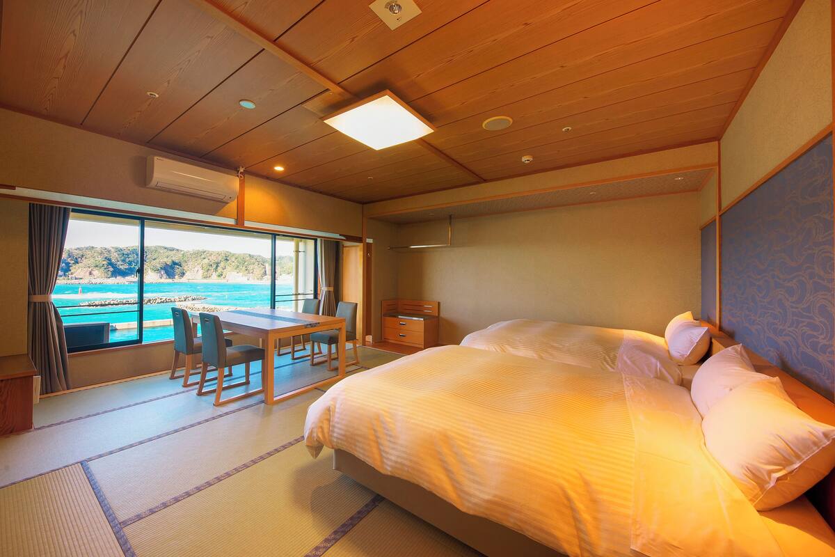 A hotel room with a bed, a dining table, chairs, a view of the sea, and a window.