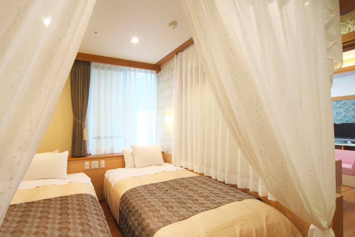 Ein Hotelzimmer mit zwei Betten, einem Fenster mit Vorhängen und einem Fernseher in der Ecke.