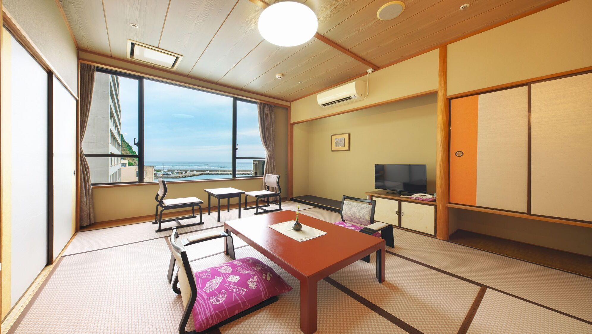 A traditional Japanese room with tatami flooring, a low table, chairs, a television, and a view of the sea.