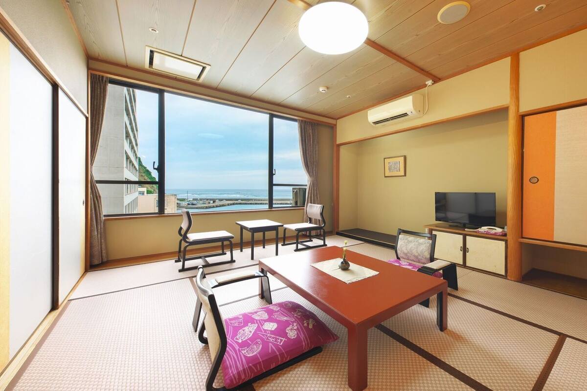 A traditional Japanese room with tatami flooring, a low table, chairs, a television, and a view of the sea.