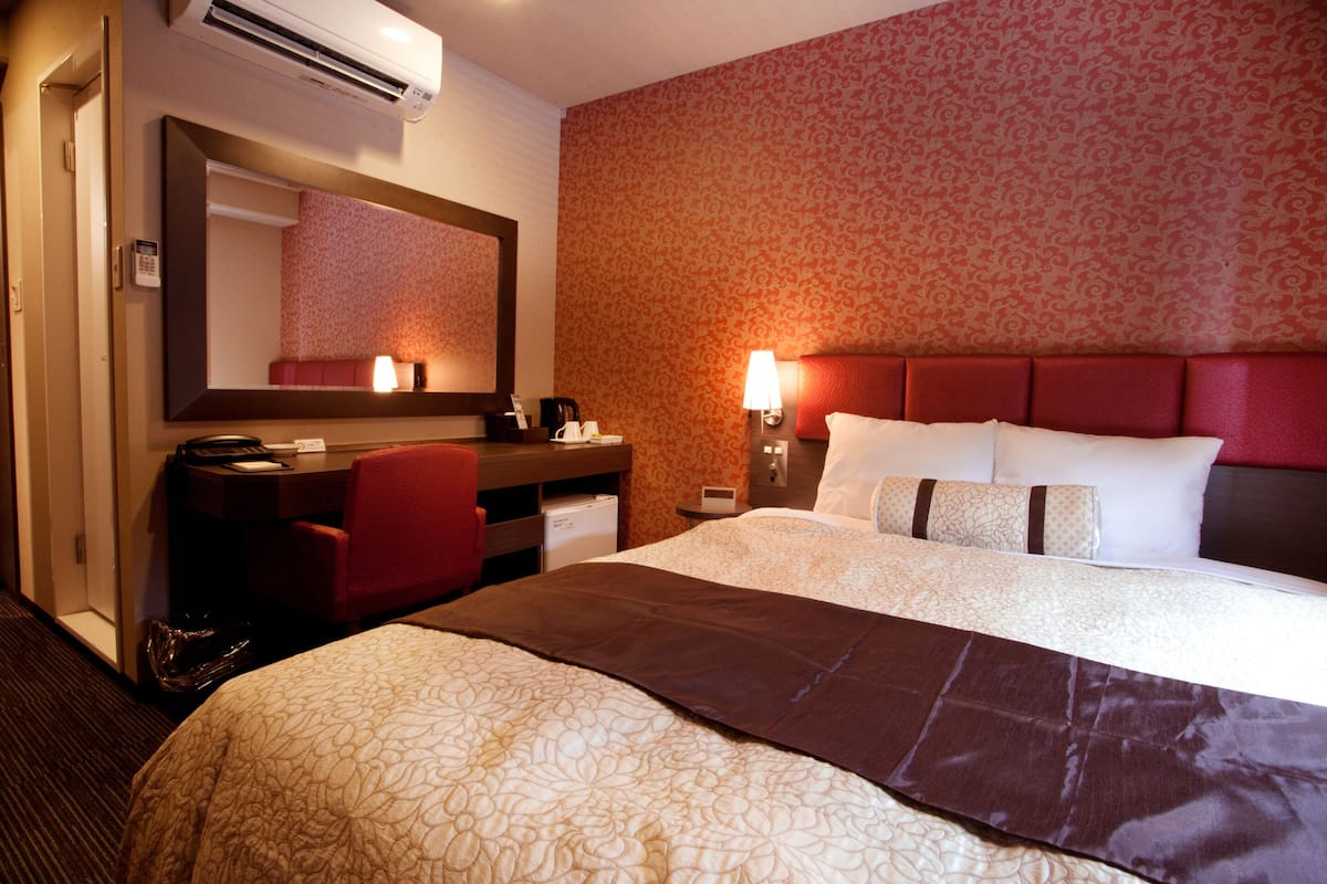 Habitación de hotel con una cama grande, un escritorio con silla, un espejo y un cabecero rojo.
