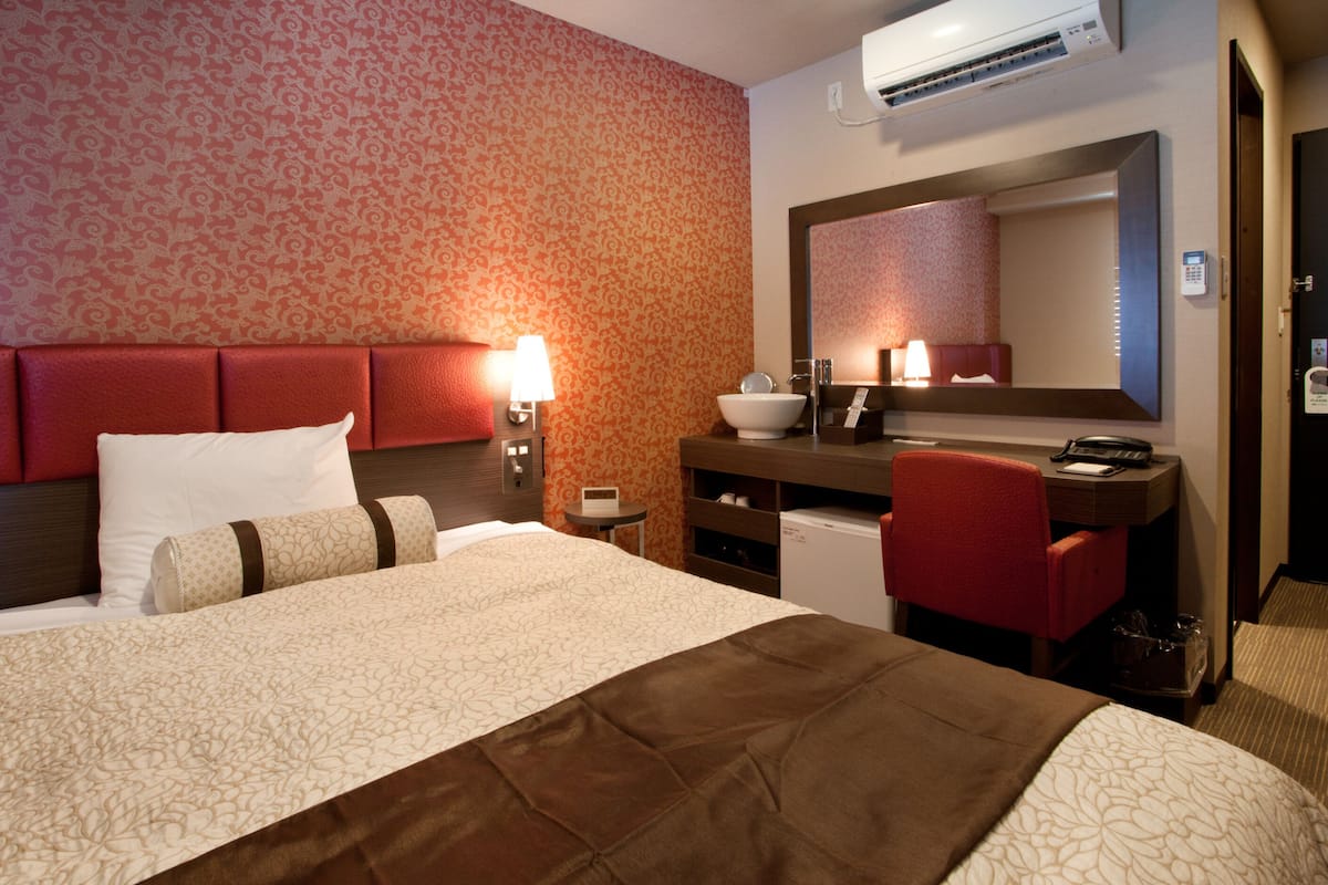 Habitación de hotel con una cama grande, un escritorio con silla, baño con lavamanos y espejo, y pared con diseño.