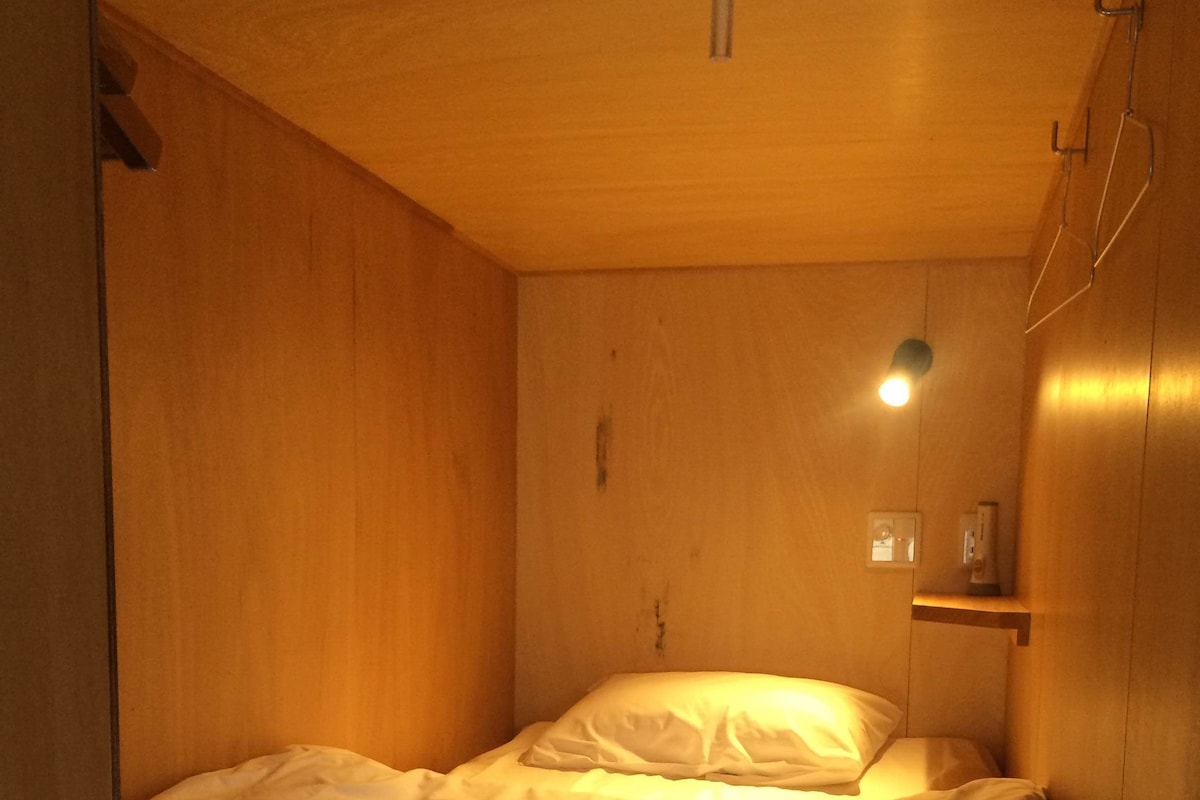 Una habitación pequeña de madera con una cama individual, una mesita de noche y una repisa en la pared.