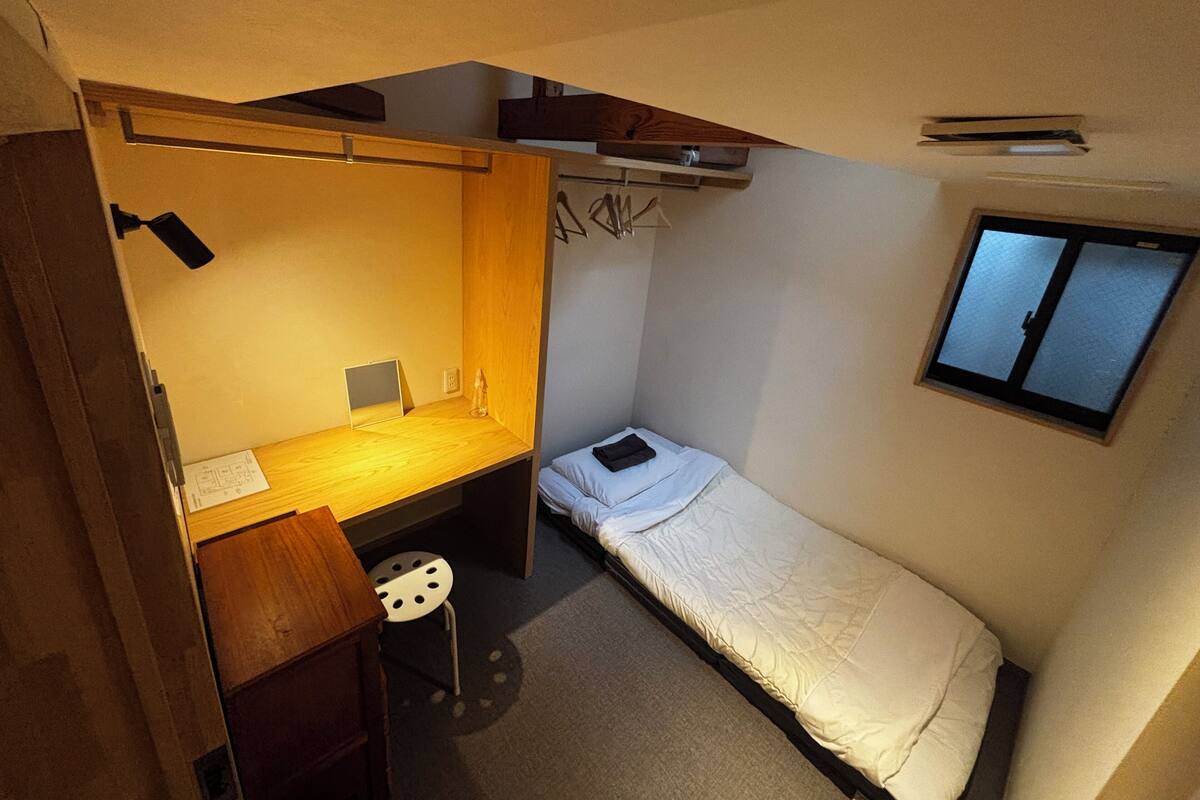 A compact dormitory room with a single bed, a desk with a chair, a small stool, and a window.
