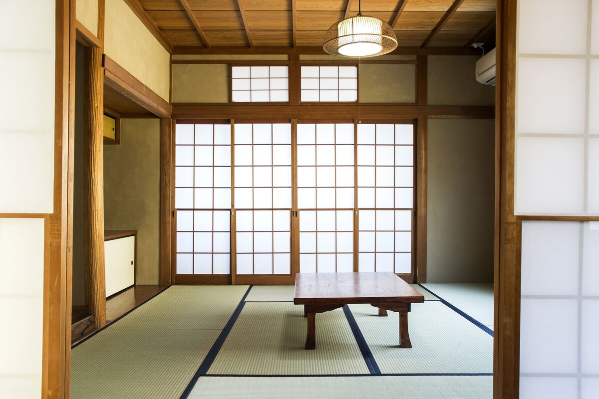 Una habitación japonesa tradicional con suelo de tatami, una mesa de madera, puertas correderas y un ventilador de techo.