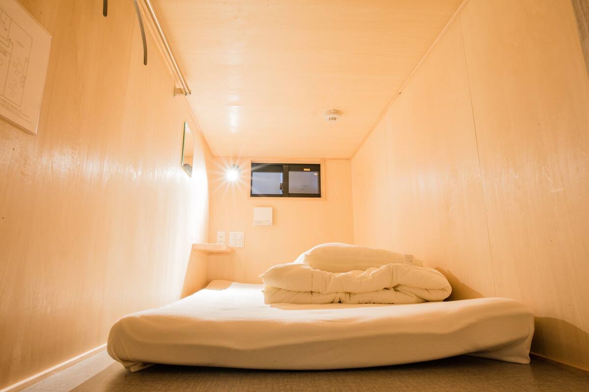 A narrow room with a bed, a small bedside table, and a wall-mounted TV.