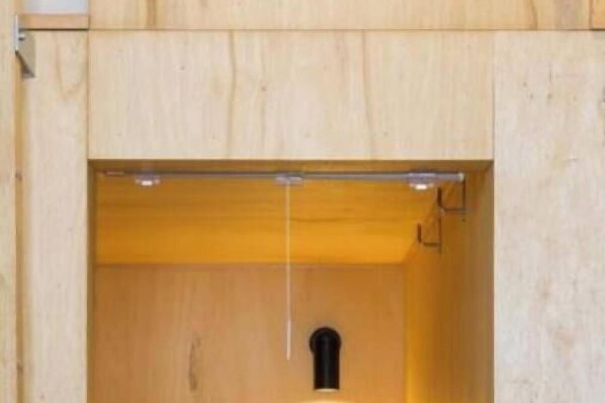 A compact wooden room with a bed, a small shelf with two items, and a wall-mounted light fixture.