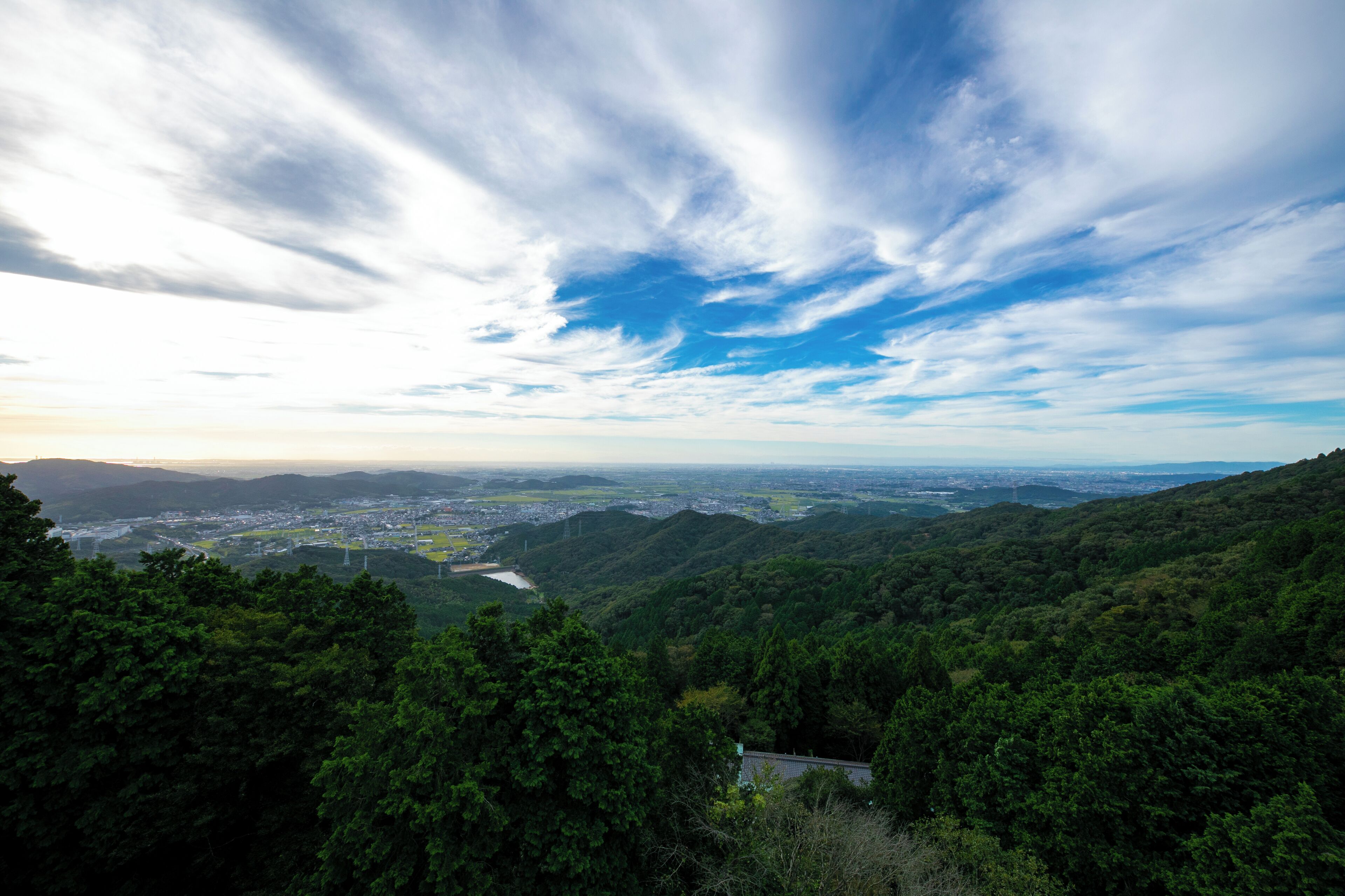 Panoramablick auf eine gebirgige Landschaft mit dichtem Wald, einem Fluss und einer fernen Stadtansicht unter teils bewölktem Himmel.