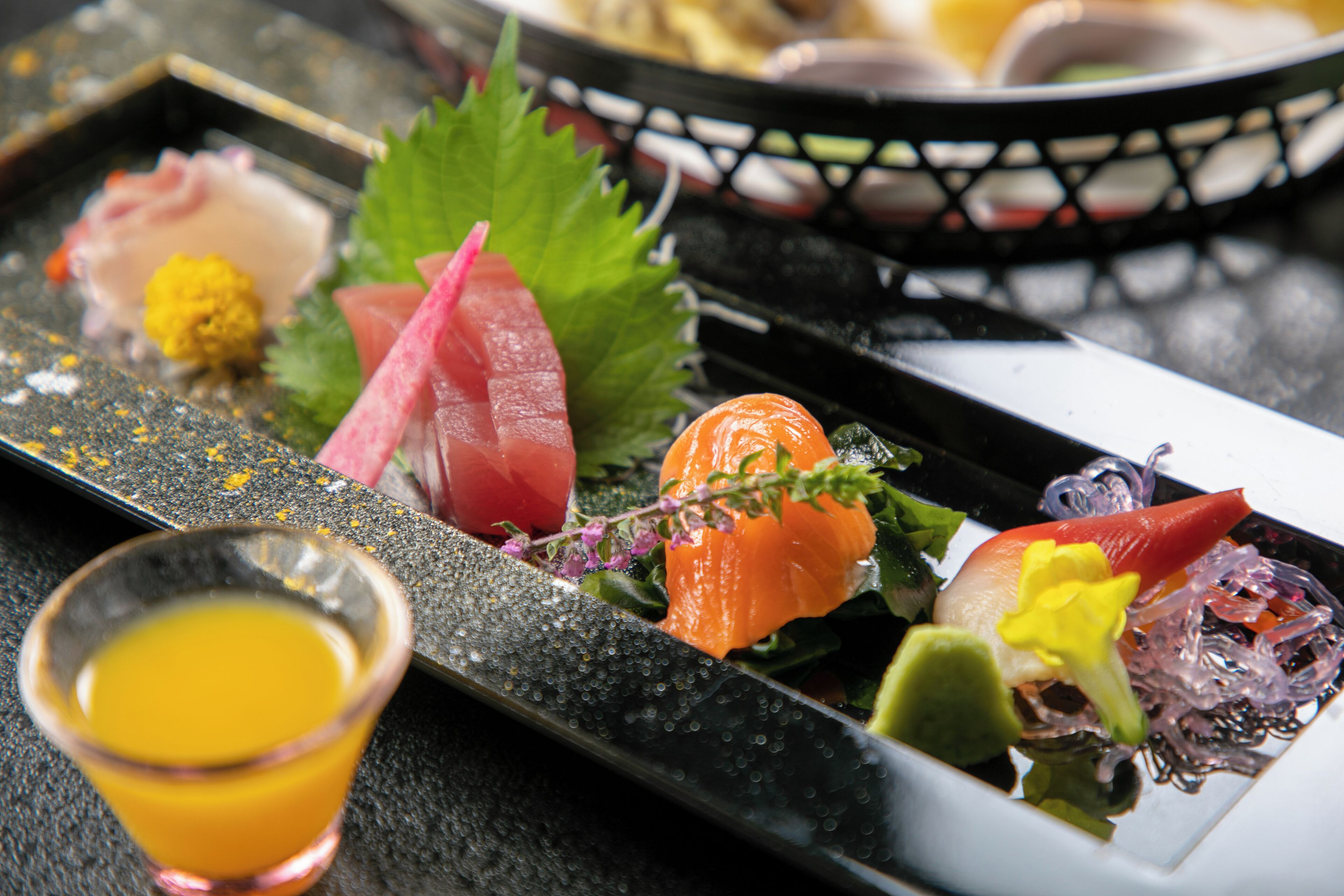 A variety of sushi dishes on a black plate with a glass of orange juice beside it.