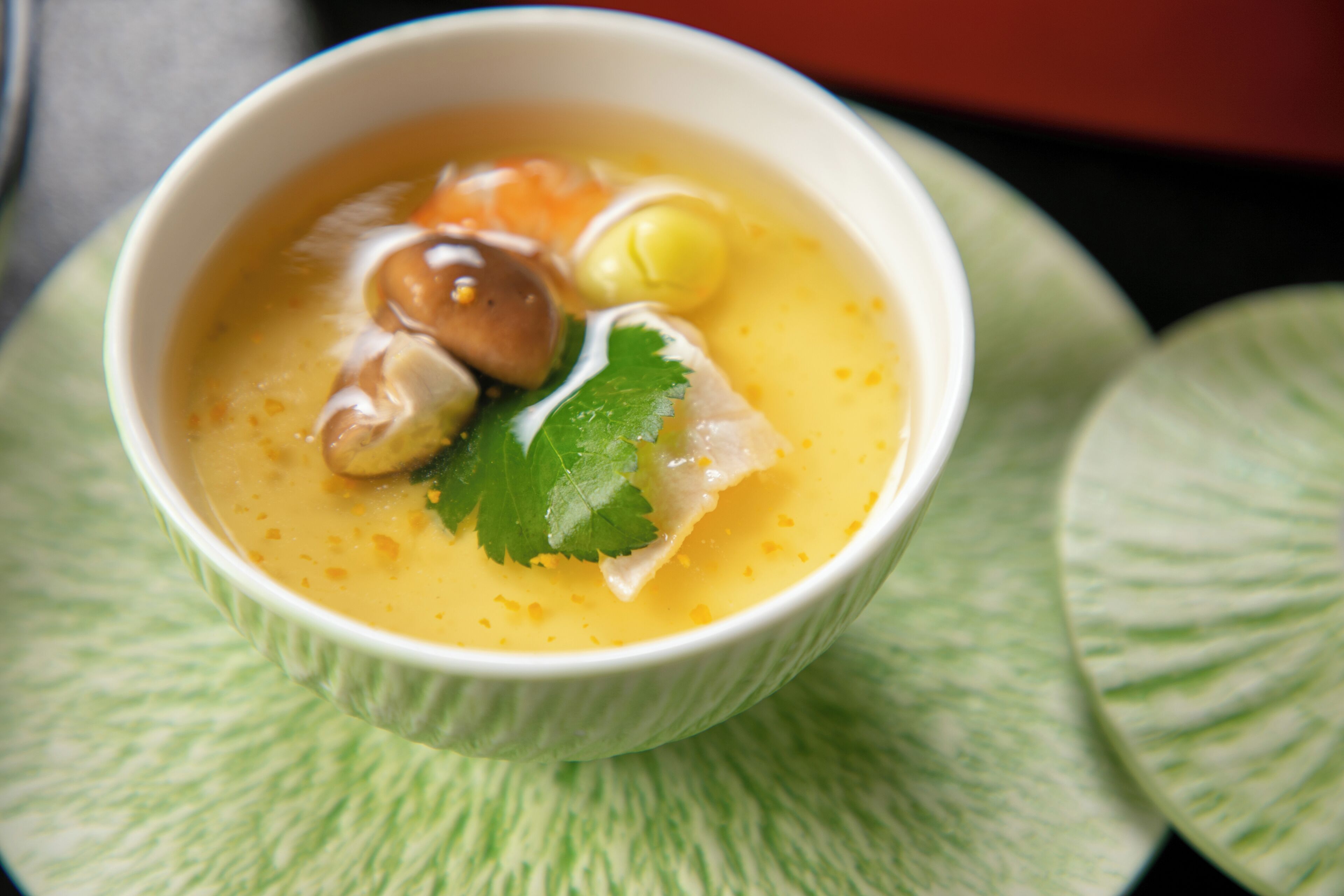 A bowl of soup with various ingredients, including mushrooms and a leafy garnish, served in a white bowl on a green textured surface.