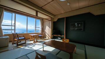 Um quarto japonês tradicional com piso de tatame, uma mesa baixa, cadeiras, uma televisão e uma grande janela com vista para o mar.