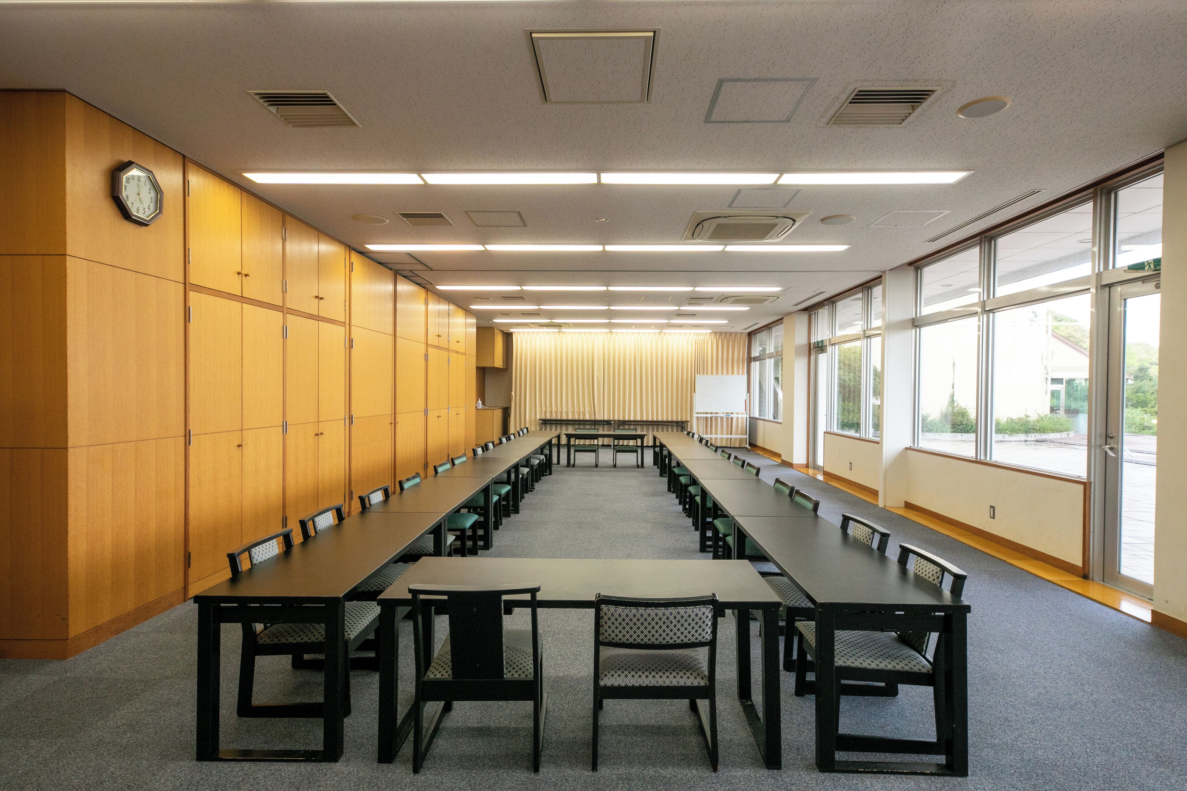 A conference room with long tables and chairs, wooden paneling on one wall, and large windows on the other.