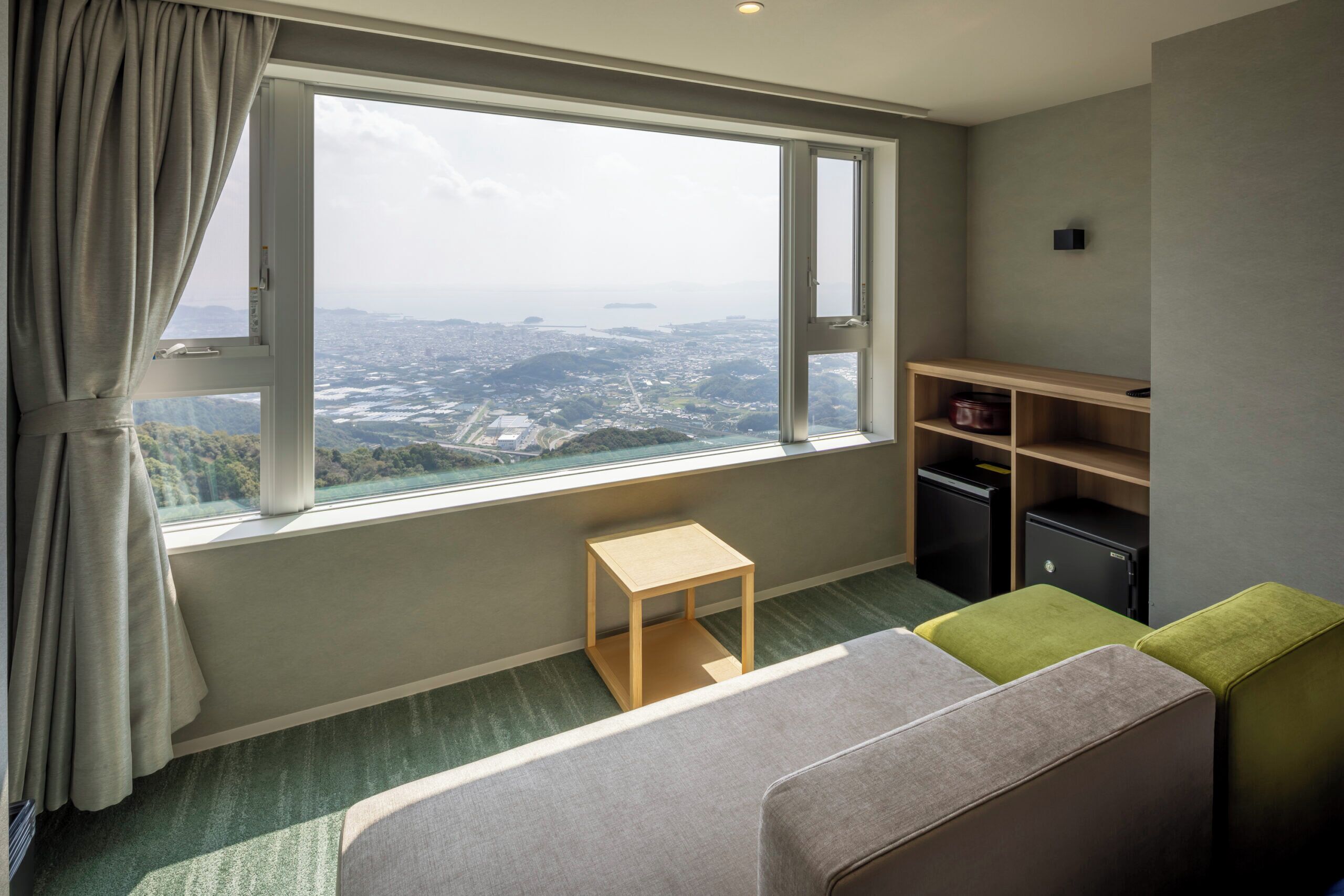 A hotel room with a large window offering a city view, a small wooden table, a green ottoman, and a shelf with storage boxes.
