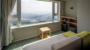 A hotel room with a large window offering a city view, a small wooden table, a green ottoman, and a shelf with storage boxes.