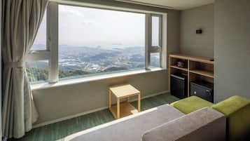 A hotel room with a large window offering a city view, a small wooden table, a green ottoman, and a shelf with storage boxes.